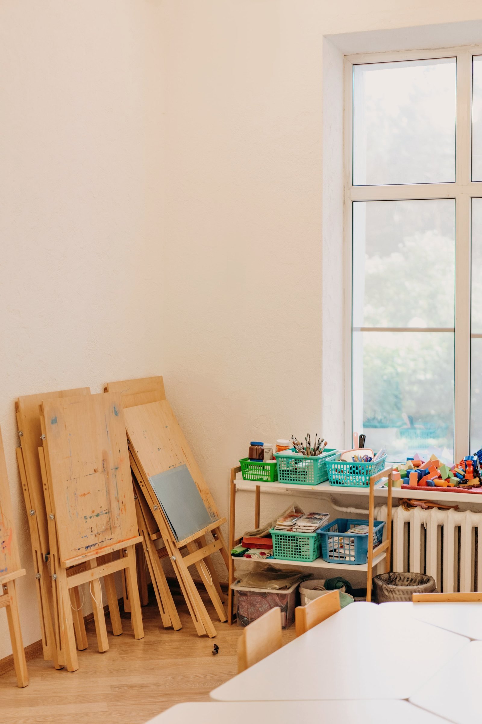Wooden art easels stacked beside baskets of brushes and paint in a bright room.